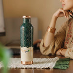Green and copper water bottle with 'pipal' branding on a table, woman sitting in the background
