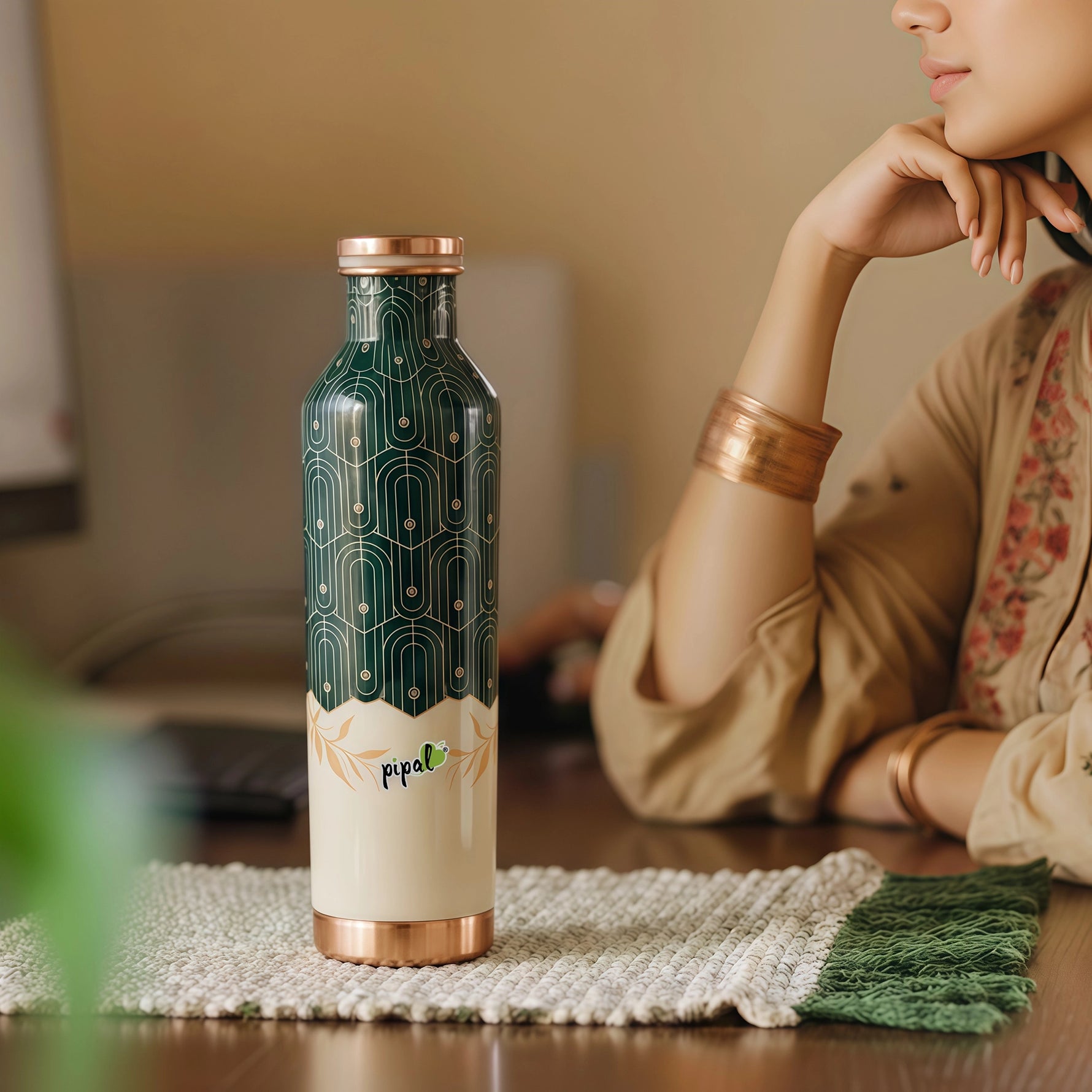 Green and copper water bottle with 'pipal' branding on a table, woman sitting in the background
