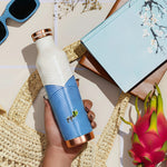 Person holding a blue and white patterned copper bottle with a straw bag, sunglasses, and dragon fruit in the background.